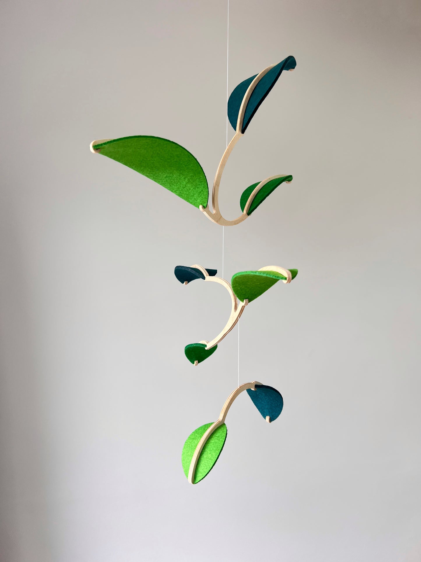 Felt Leaf Mobile 

   Elevate your space with this versatile Midcentury Modern Mobile, designed for both nurseries and adult living areas. Crafted from natural wood with deep teal and green shades of soft felt, this handmade nursery mobile embodies a minimalist and Scandinavian aesthetic. Its unique geometric design creates a stunning hanging sculpture that fits seamlessly into nursery decor and neutral relaxing spaces. 